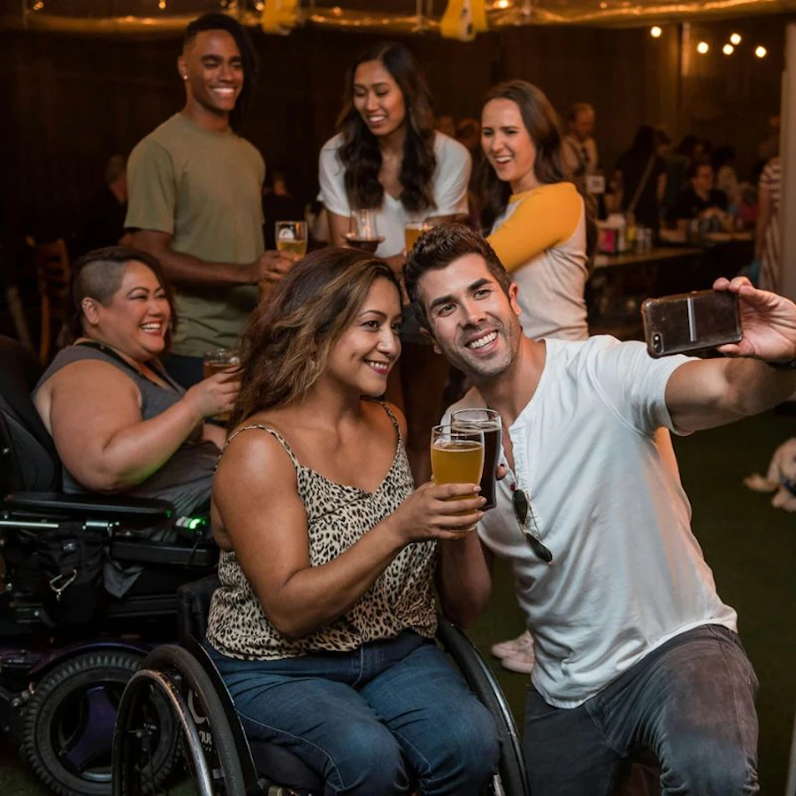 A diverse group of friends at a social gathering, smiling and posing for a selfie. One woman is in a wheelchair, holding a beer glass, while a man beside her takes the photo. Others in the background are also enjoying their drinks and the company. The setting appears to be a warm and welcoming party.